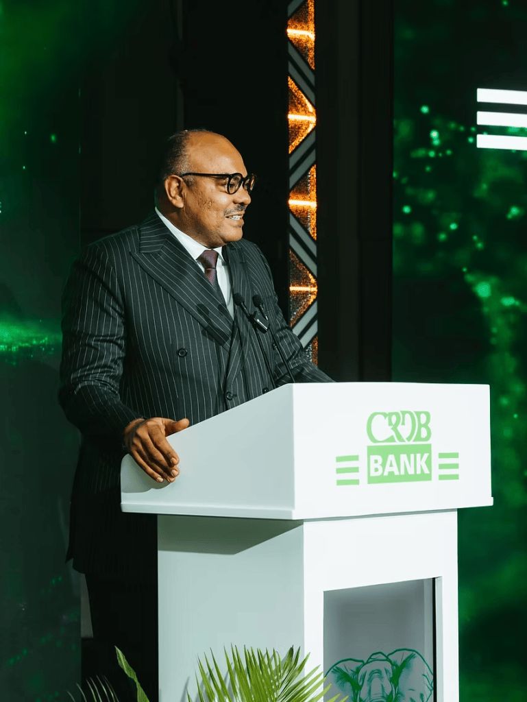 A speaker in a pinstriped suit standing behind a podium with the logo of CRDB Bank, addressing an audience in a well-lit venue with green-themed decor.