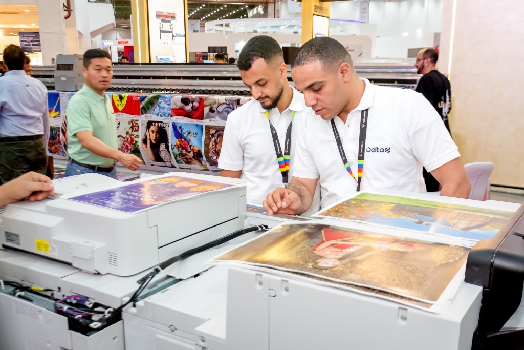 Two professionals in white shirts examining printed images on a digital printer at a trade show, with colorful printed materials displayed in the background.
