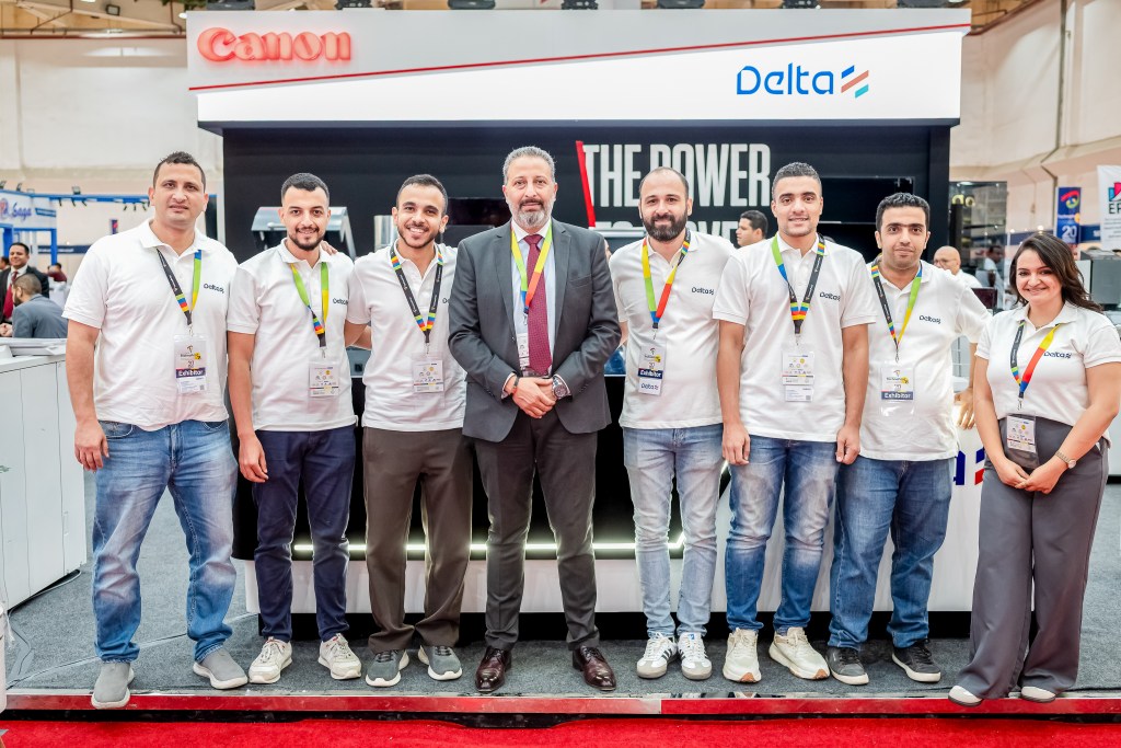 Group of seven individuals posing together at a trade show, with Canon and Delta logos in the background. All are wearing matching white shirts and name tags.