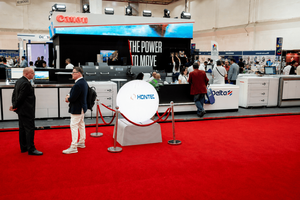 Trade show scene featuring a Canon booth with the phrase 'THE POWER TO MOVE' displayed, red carpet, and attendees engaging with various exhibits in the background.