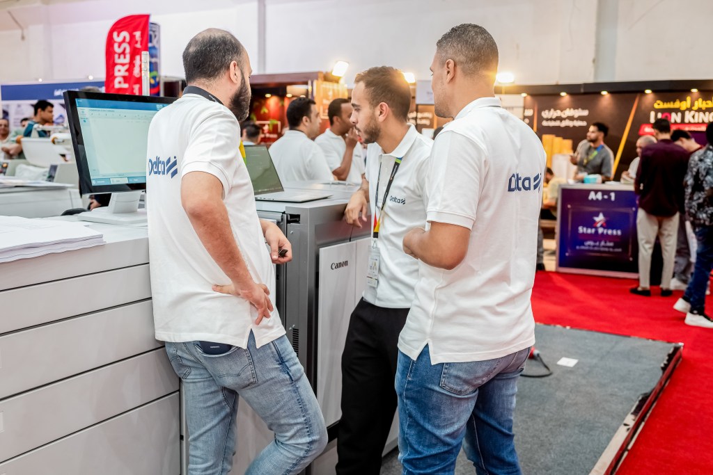 Three professionals in white shirts discussing around a computer at a trade show booth, with people and displays in the background.
