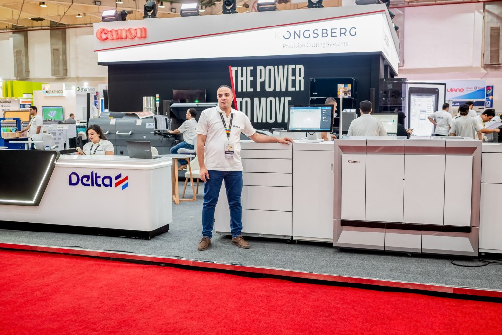 A trade show booth featuring Canon equipment and the Delta logo, with several employees interacting with technology. A man stands in front of a large Canon machine while others work in the background.