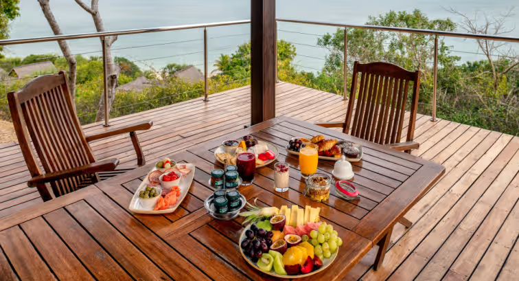 A beautifully arranged breakfast spread on a wooden table, featuring fresh fruits, pastries, juices, and yogurt, with a scenic view of the ocean and greenery in the background.